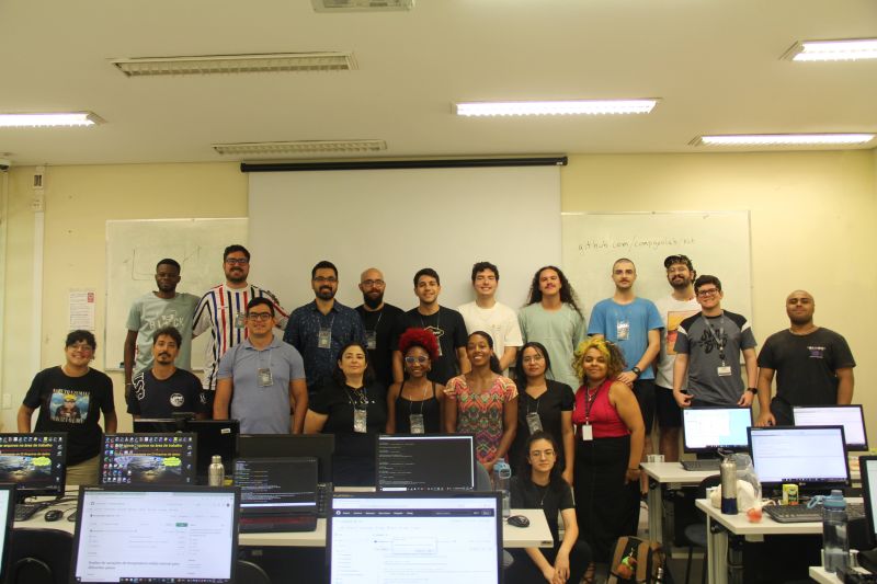 A diverse group of individuals stands in a classroom setting, with computers and workstations in the foreground.