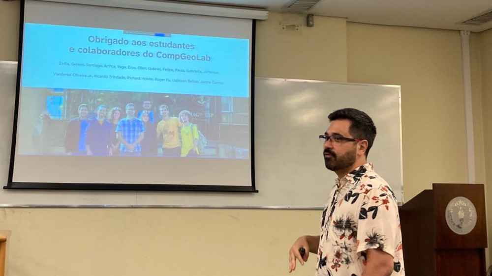 Photo of a man with glasses, short dark hair and a short beard wearing a flower patterned shirt in the middle of speaking and projected slides in the background saying 'Obrigado aos estudantes e colaboradores do CompGeoLab' with a photo of a group of people.